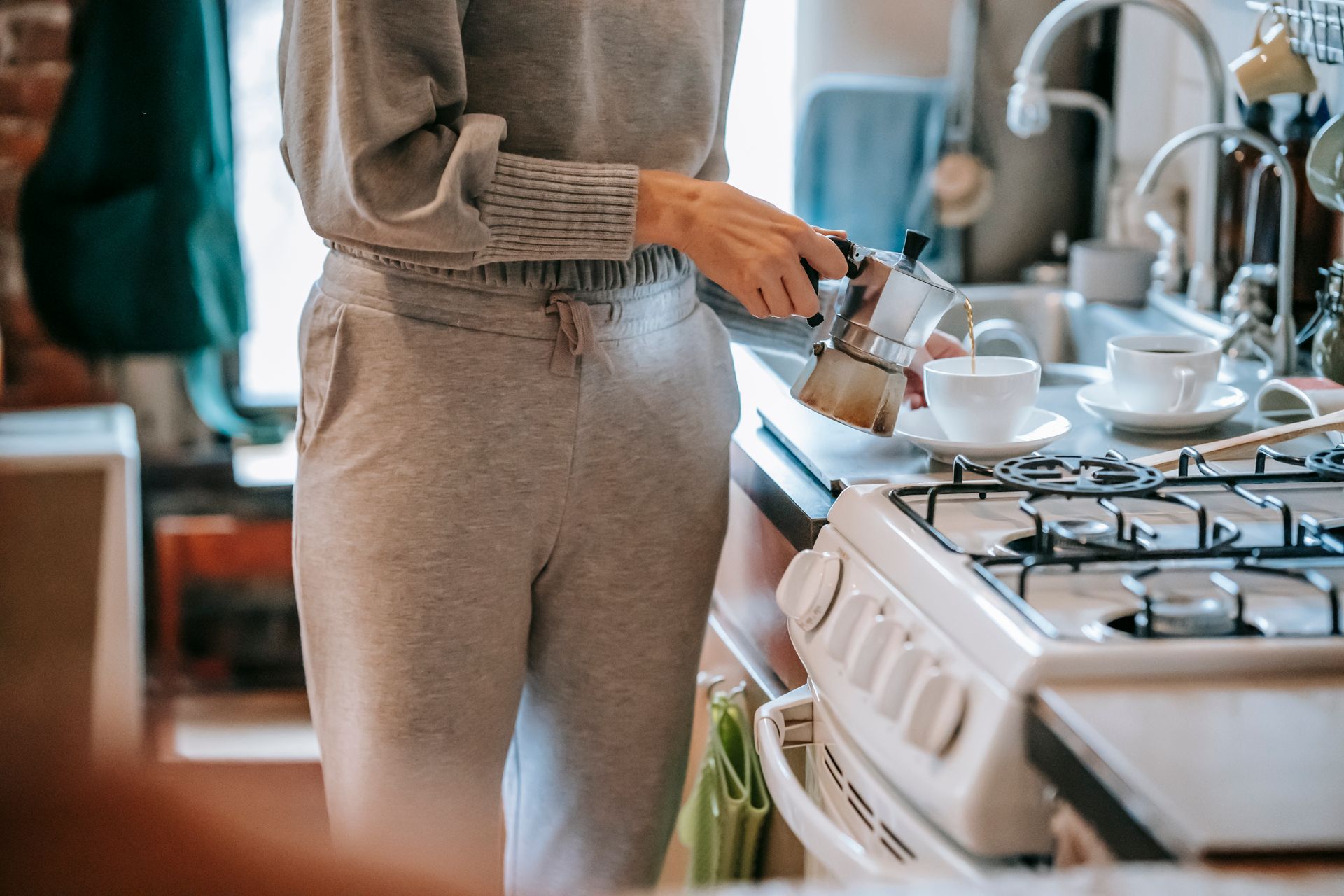 A woman pouring freshly brewed coffee using a moka pot in a cozy kitchen environment.