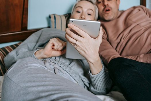 Crop adult diverse married couple in casual clothes cuddling while lying on bed and watching interesting movie on tablet