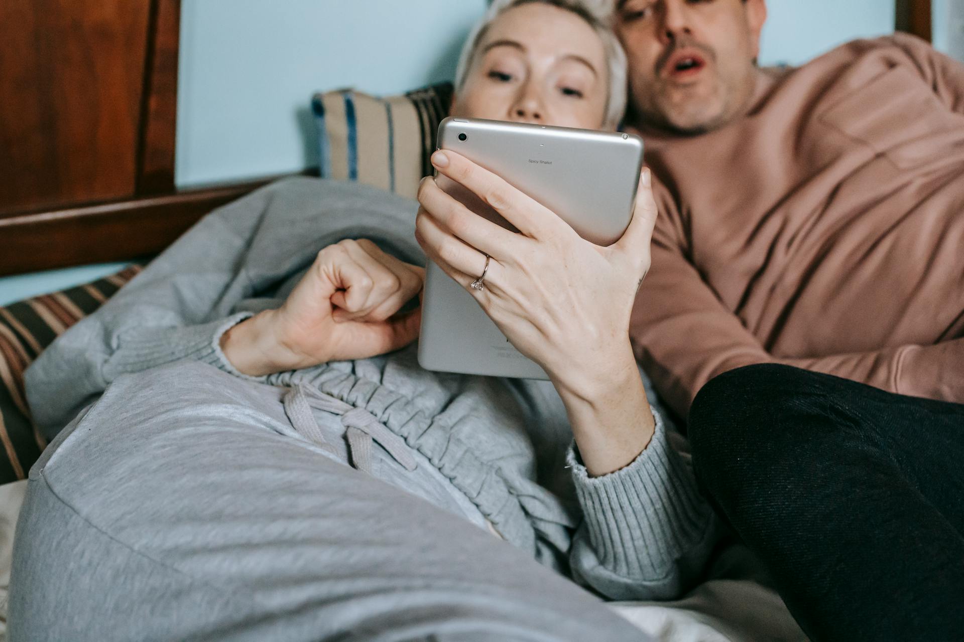 Crop adult diverse married couple in casual clothes cuddling while lying on bed and watching interesting movie on tablet