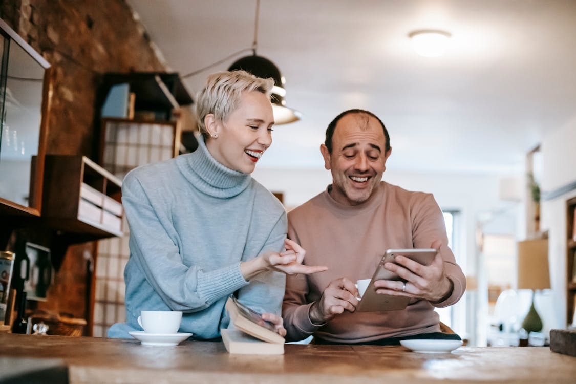 Free Cheerful adult diverse couple in casual clothes watching funny video on tablet and laughing while drinking coffee at table in apartment Stock Photo