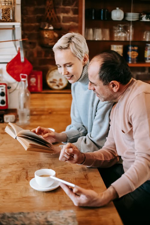 Free Side view of curious middle aged ethnic man looking at book reading by wife while sitting together at wooden table and drinking coffee during breakfast at home together Stock Photo