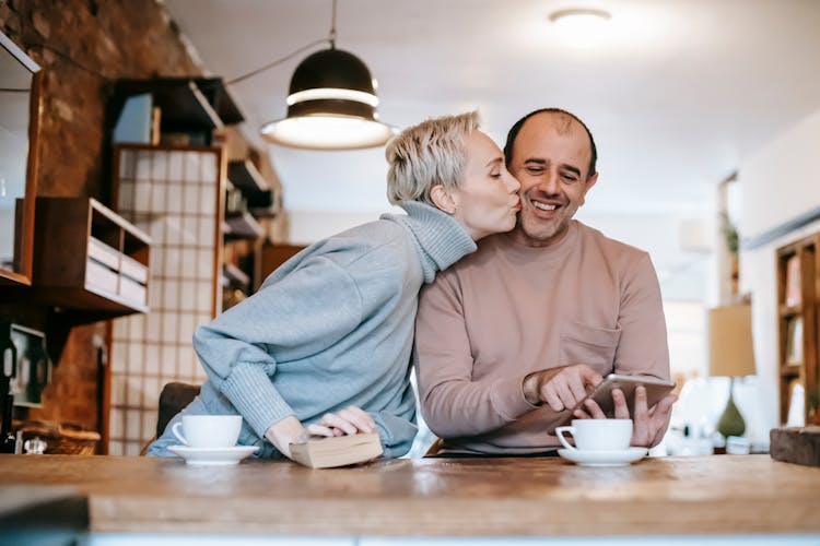 Happy Woman Kissing Cheek Of Ethnic Husband Surfing Tablet At Table In Kitchen