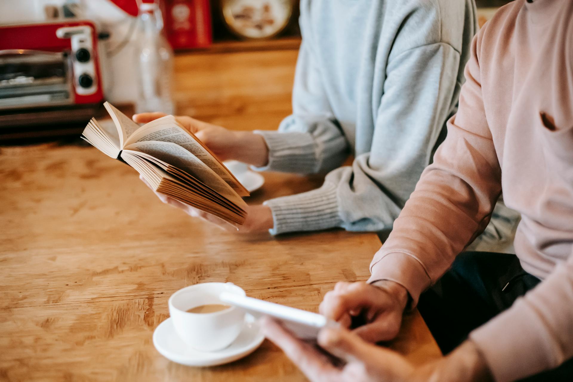 From above of crop faceless spouses in casual clothes using tablet and reading book while sitting at wooden table with cups of cappuccino at home