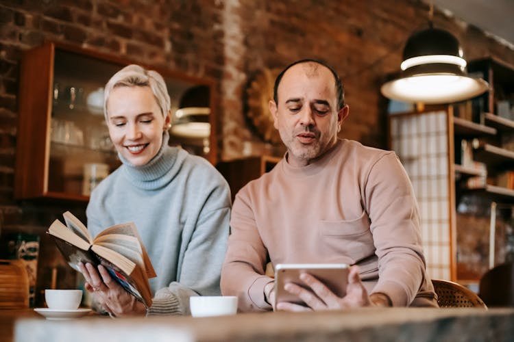 Adult Multiracial Spouses Sitting At Table While Using Tablet And Reading Book At Hone