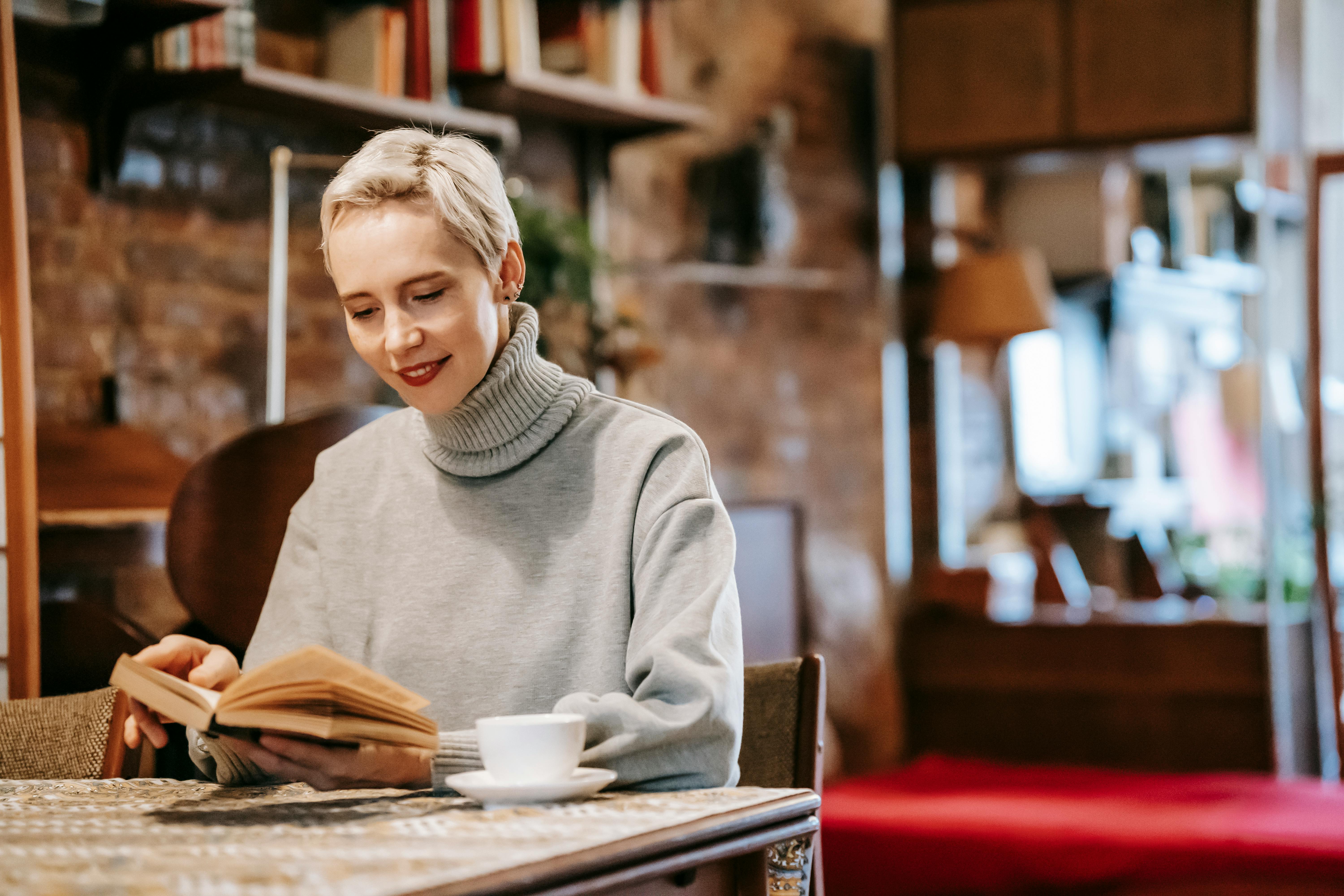 smiling woman reading novel at table and drinking coffee