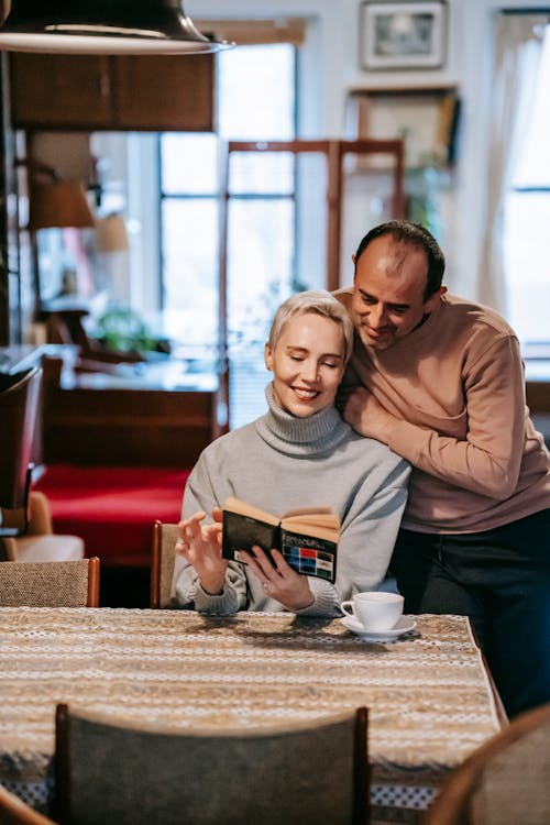 Positive adult multiethnic couple reading book at table in apartment