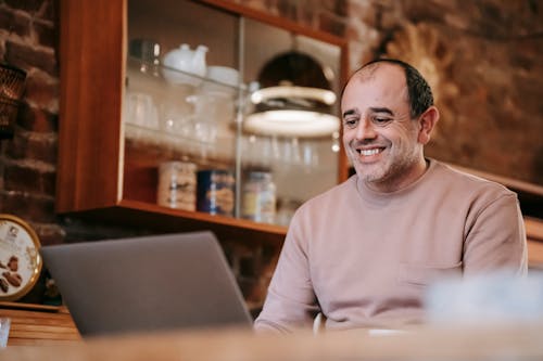 Free Content adult ethnic male freelancer in casual clothes smiling while working online on laptop sitting at table at home Stock Photo