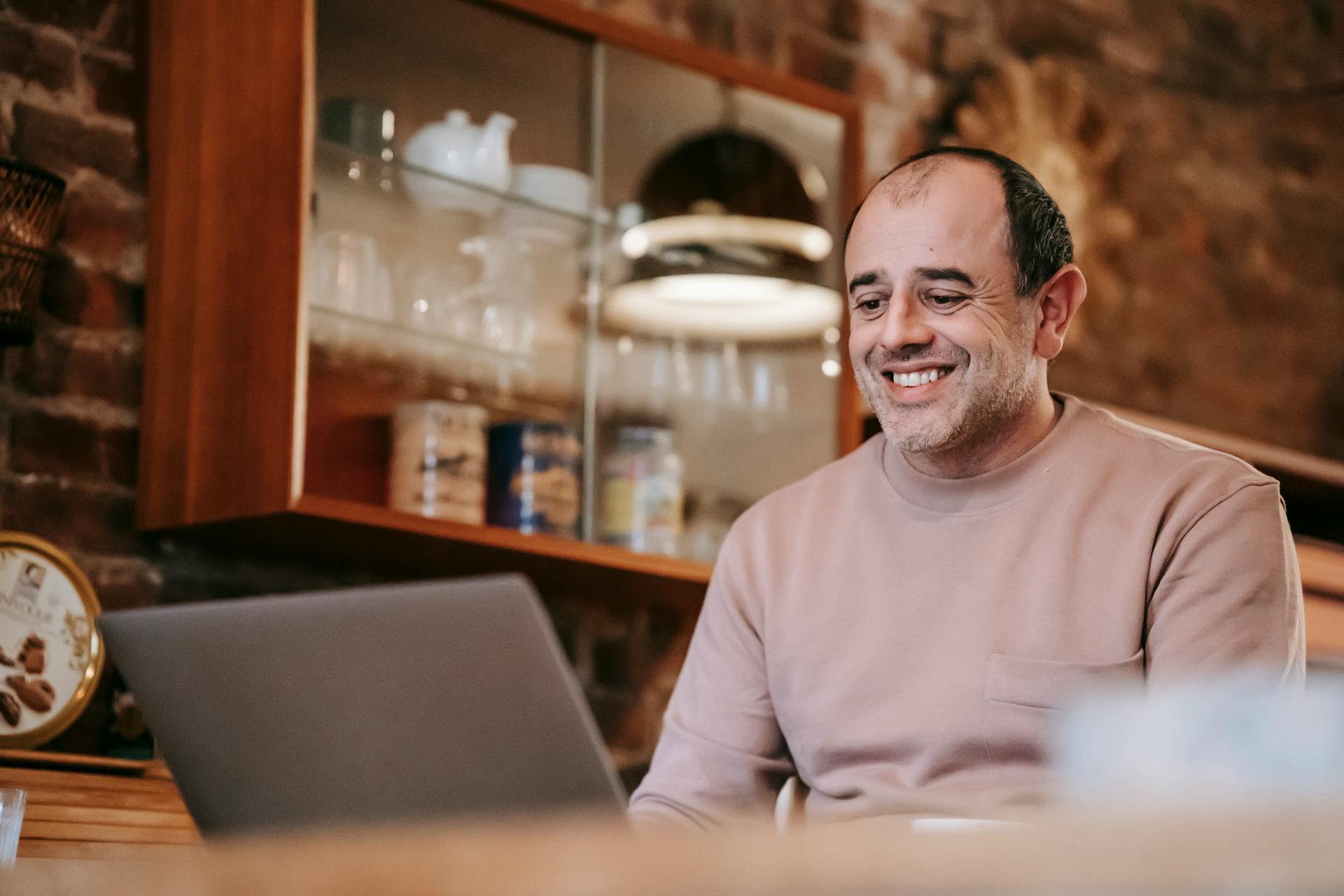 Content adult ethnic male freelancer in casual clothes smiling while working online on laptop sitting at table at home