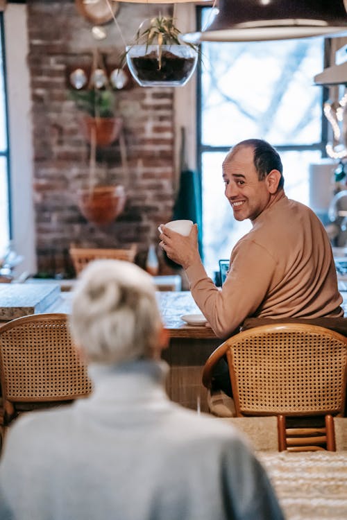 Cheerful ethnic male drinking coffee and communicating with unrecognizable wife at home