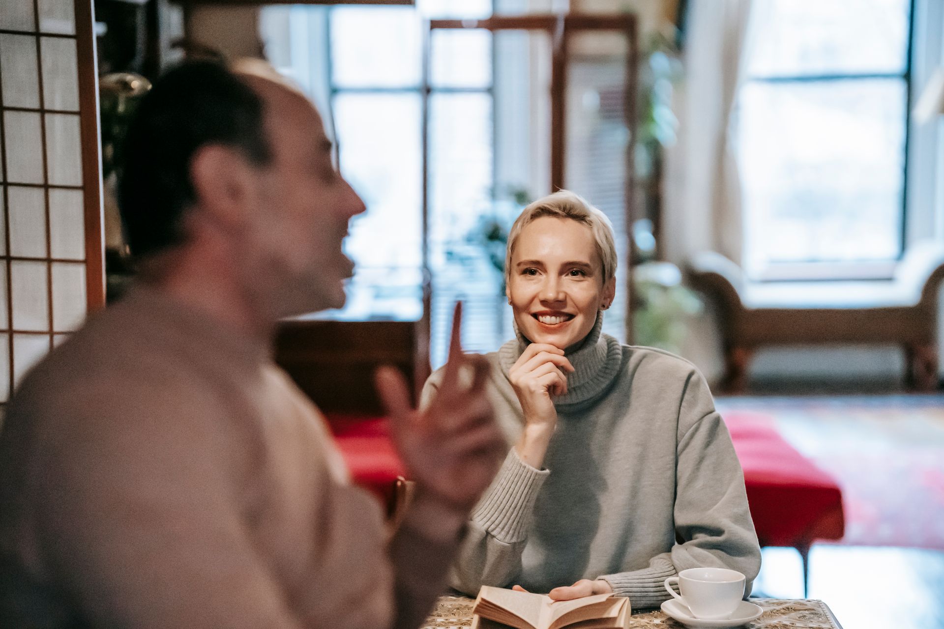 Optimistic adult lady sitting at table with book and touching chin while communicating with unrecognizable husband during weekend at home