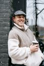 Side view of smiling adult male in warm clothes with takeaway beverage looking at camera while leaning on brick wall