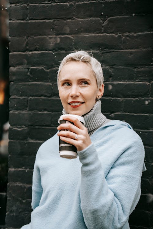 Adult contemplative female in warm clothes with takeaway hot drink looking away against brick wall