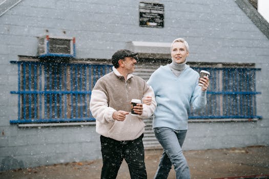 Adult couple with hot drinks to go talking while strolling against old house during snowfall in town