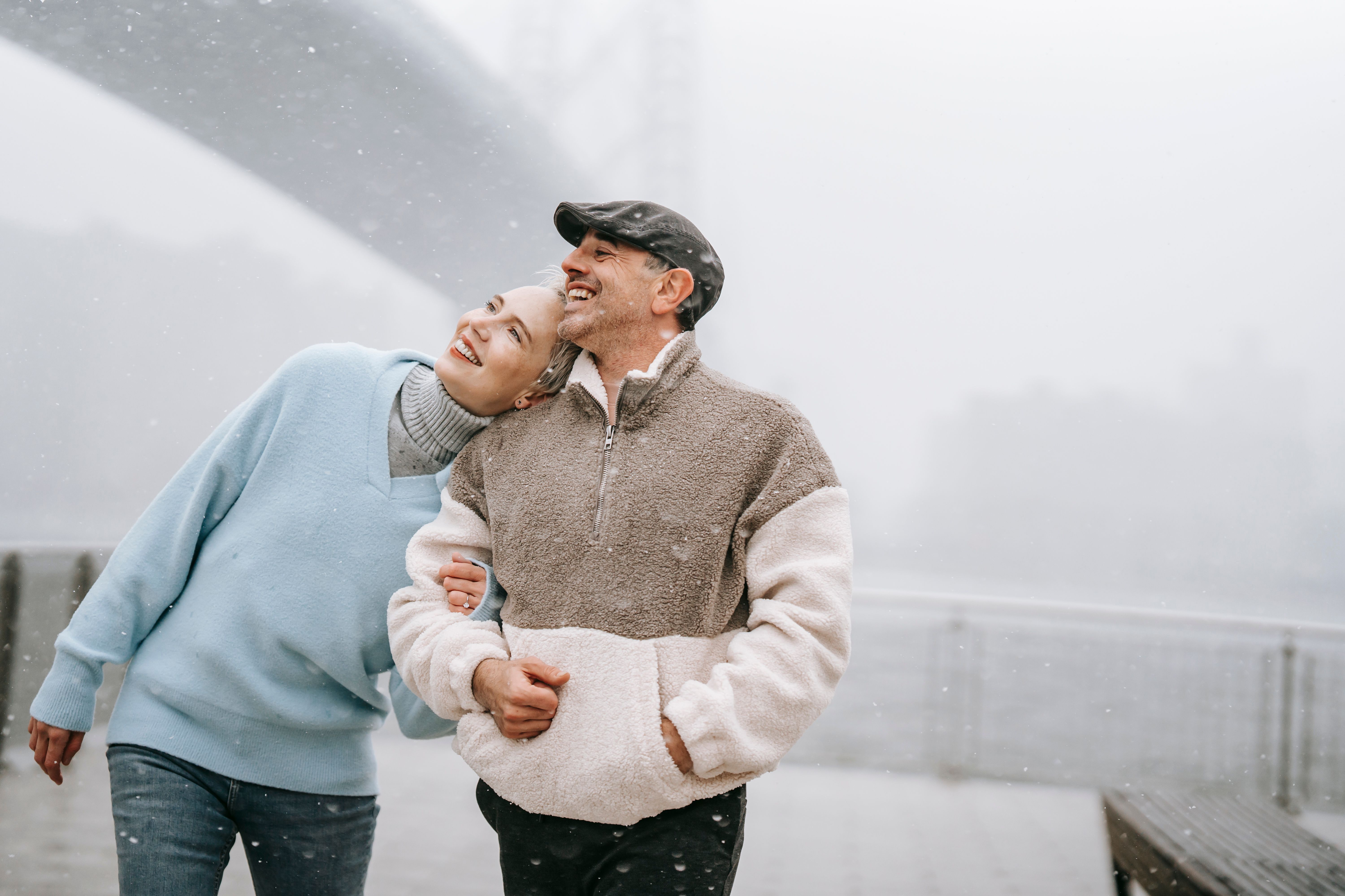 Smiling woman leaning on shoulder of male partner while enjoying snow and looking up in town on foggy day