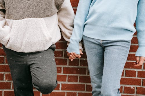 Crop unrecognizable couple in warm sweaters standing near brick wall of building and holding hands in snowy day