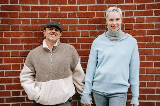 A cheerful couple holds hands outside against a red brick wall, showcasing happiness.