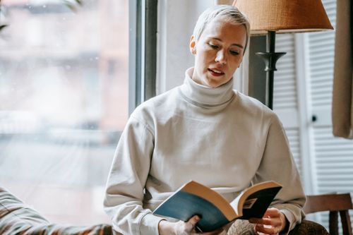 Free Intelligent content female reading volume of fascinating book near window of lounge in daytime Stock Photo