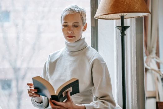 Focused smart female with short hair reading interesting book near lamp and window in daytime