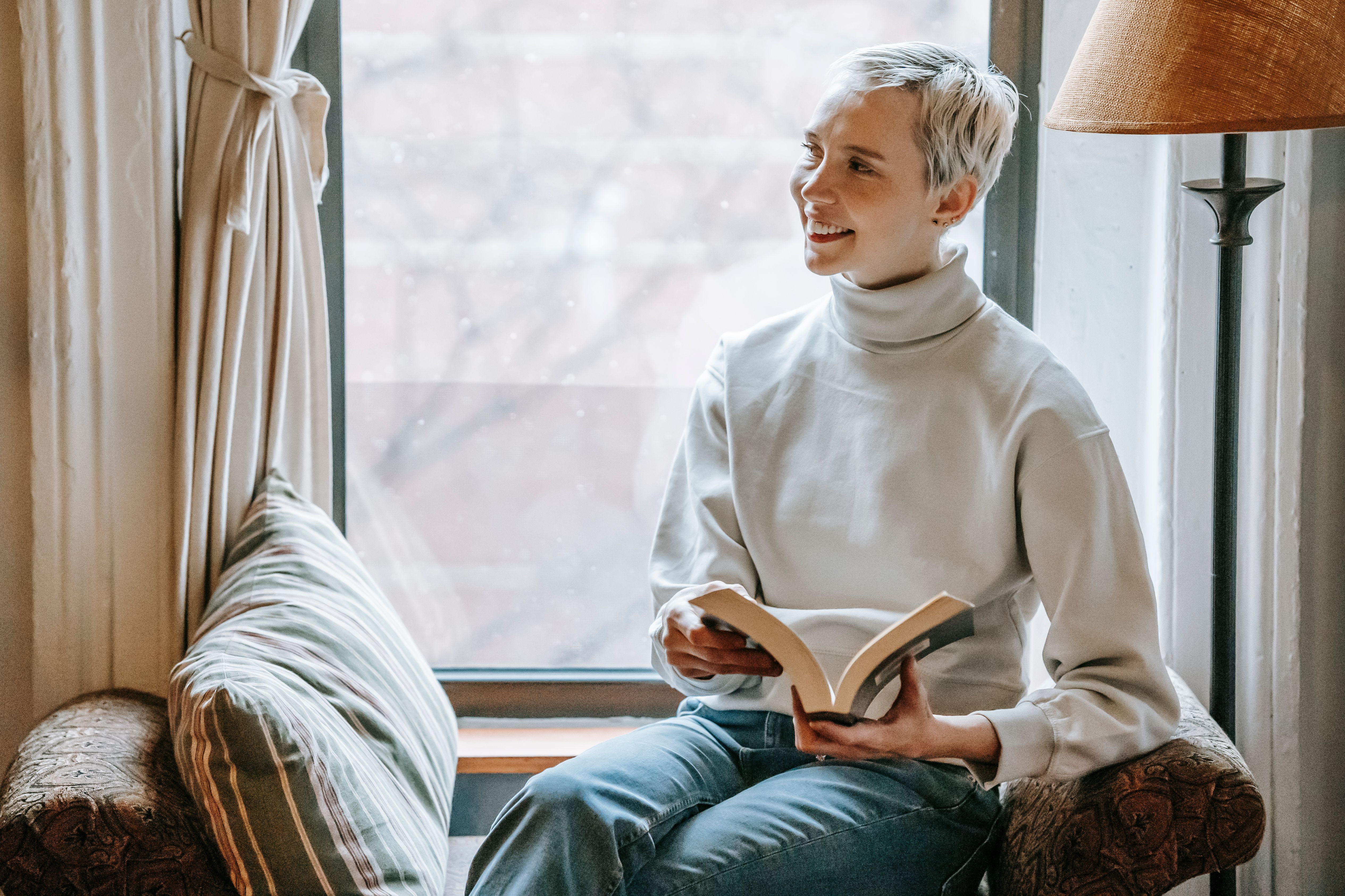 Cheerful female with interesting fiction resting on soft sofa and smiling while looking away