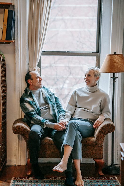 Full body of cheerful middle aged couple smiling and talking near window in cozy living room