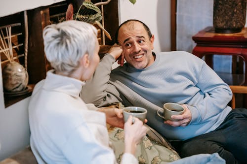 Positive diverse man and woman in casual clothes drinking cups of tea and talking while spending time in cafe in daytime