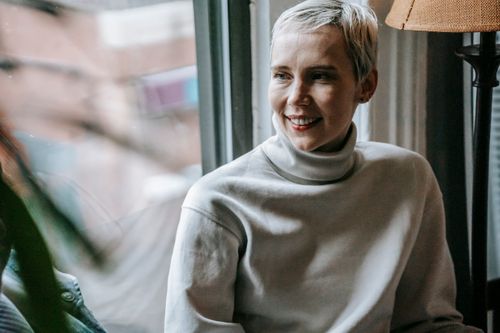 Free Smiling woman in casual clothes and red lipstick looking away near window and plant with green leaves near floor lamp in light room Stock Photo