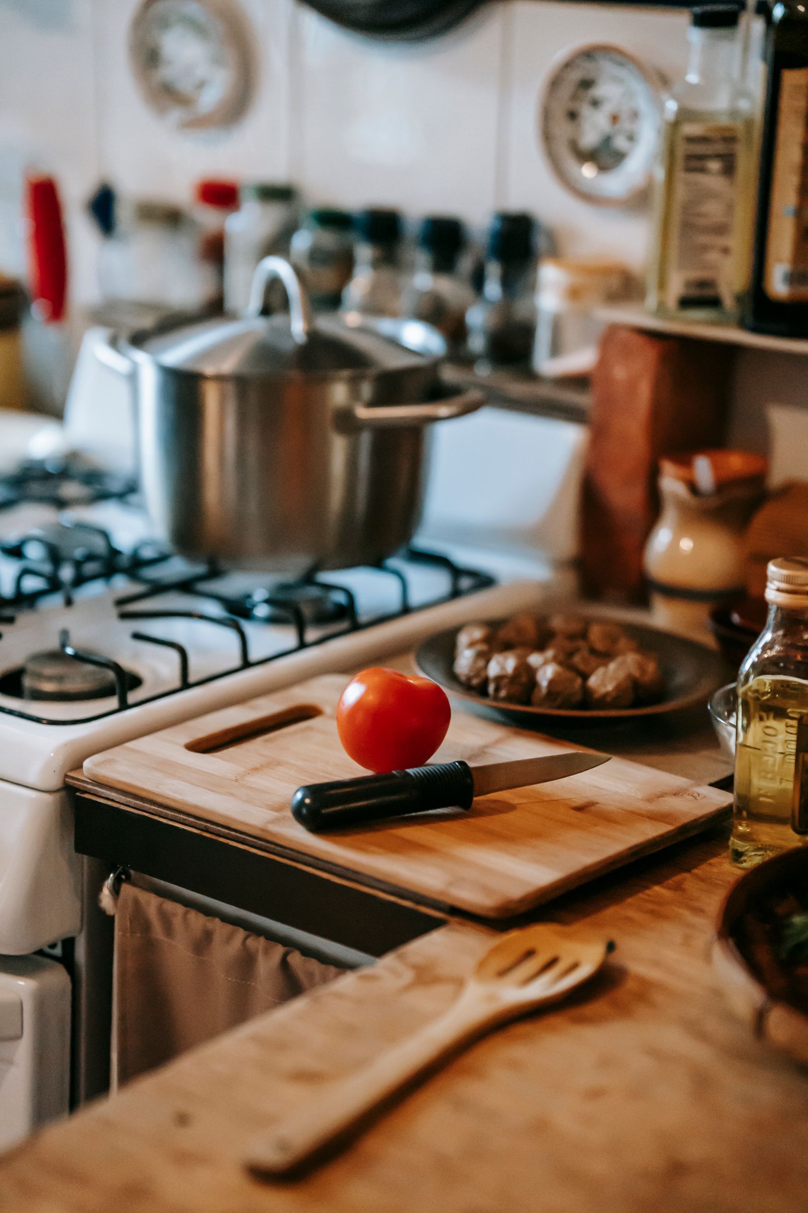 Kitchen interior with counter with tomato and knife on wooden chopping board near plate with meat and various glass bottles near stove with pan near wooden spoon