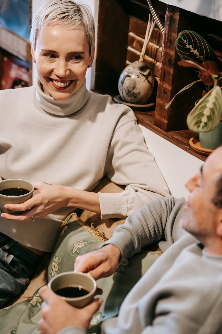 Smiling Couple On Sofa With Mugs With Coffee In Cafe