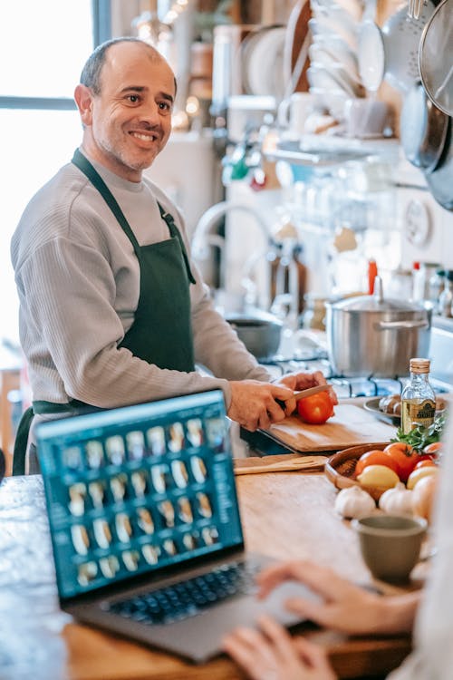 Smiling man cutting tomato against crop partner with laptop