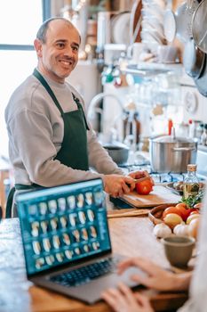 Cheerful male with tomato on chopping board interacting with crop anonymous friend with photo gallery on netbook screen in kitchen