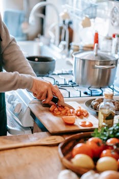 Crop unrecognizable person cutting ripe tomato with knife on chopping board against gas stove at home