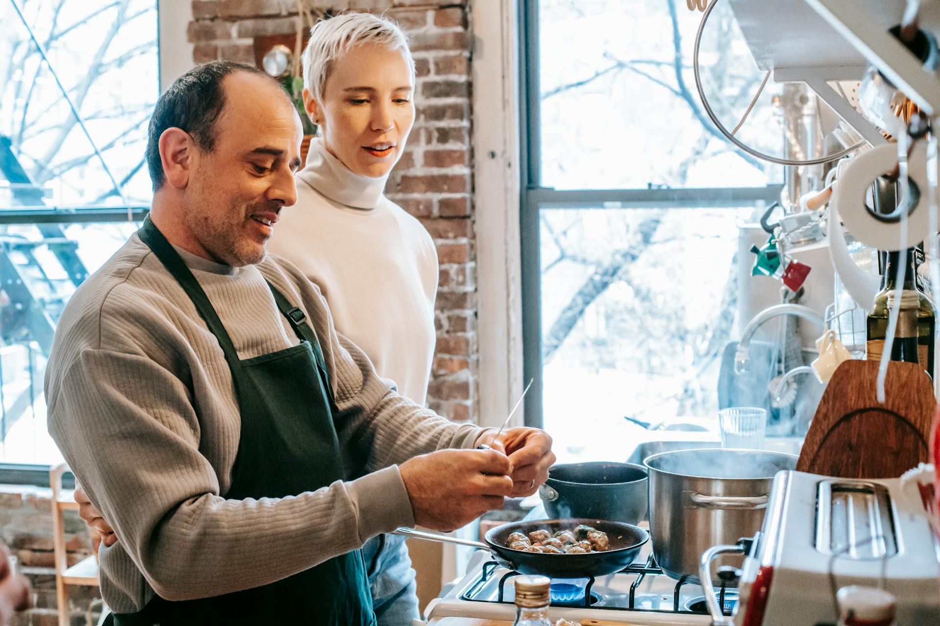 Smiling man in apron cooking against attentive girlfriend and gas stove with meatballs in pan at home