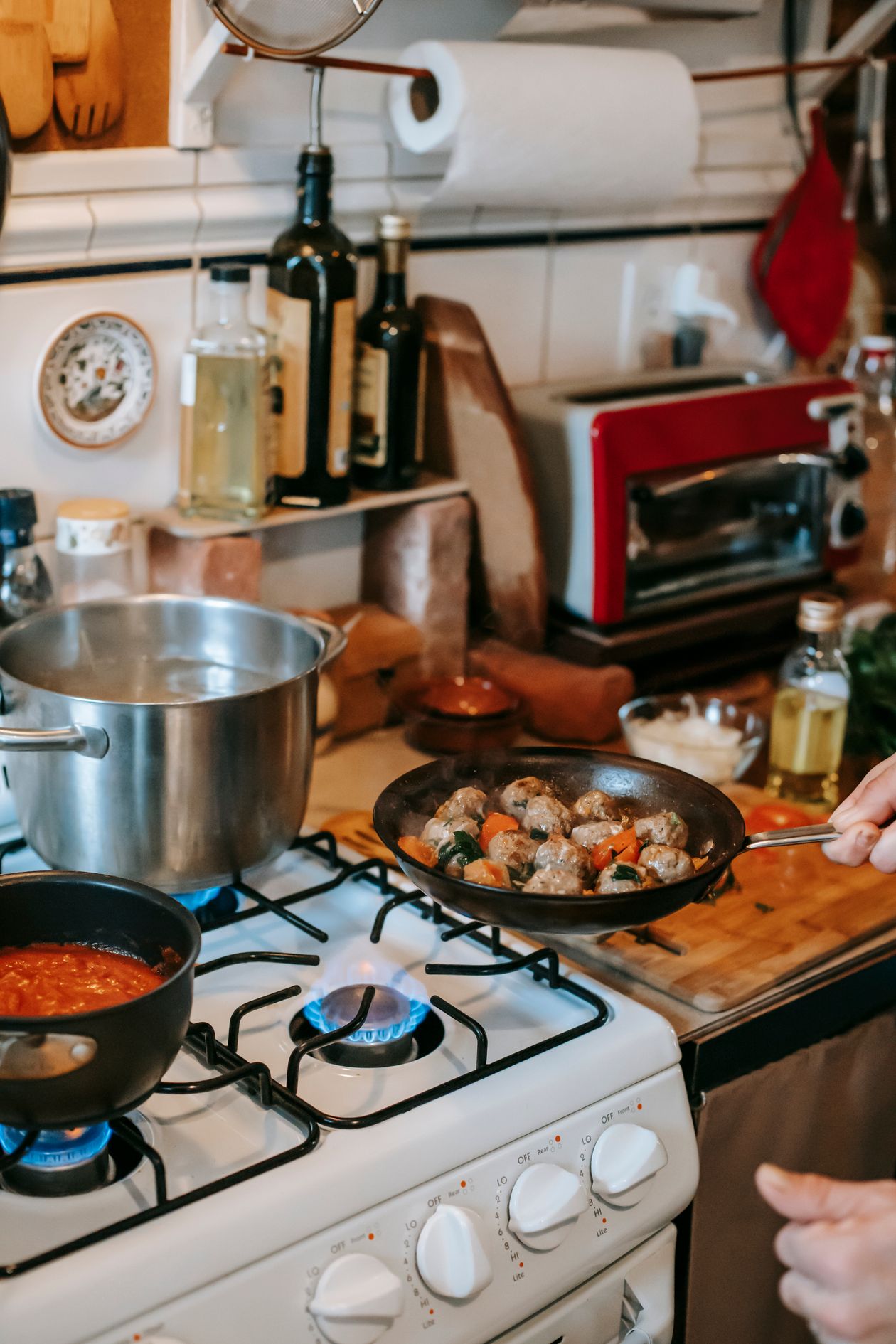Home cook holding a pan with chicken meatballs cooking on a gas stove with saucepans in the background