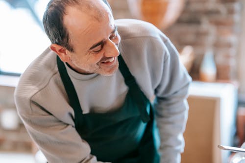 Crop smiling male in apron leaning forward during cooking process in house on blurred background