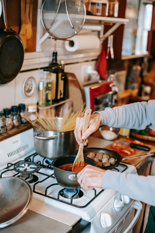 Crop person stirring tomato sauce against meatballs in kitchen