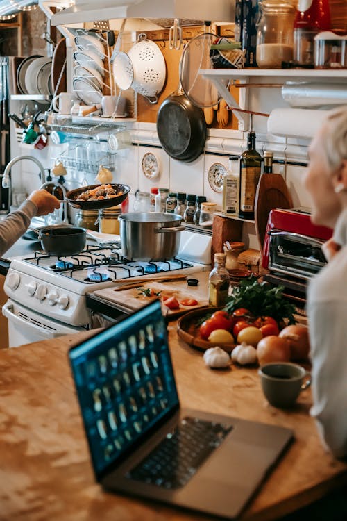 Free Side view of crop unrecognizable woman smiling and working remotely on laptop sitting at table in kitchen near anonymous husband preparing dinner Stock Photo