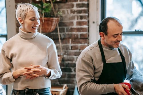 Free Positive adult woman in casual clothes smiling and looking at ethnic husband in apron preparing delicious dinner in kitchen Stock Photo