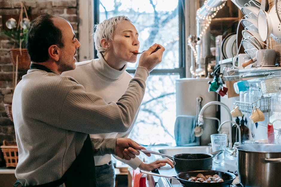 Side view of content ethnic man in casual clothes and apron feeding happy wife while preparing delicious dinner at stove in kitchen