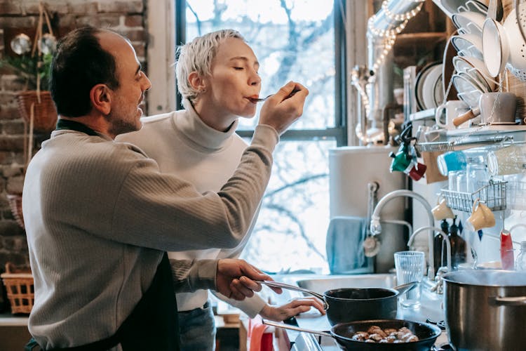 Excited Woman Tasting Food Cooked By Ethnic Husband In Kitchen