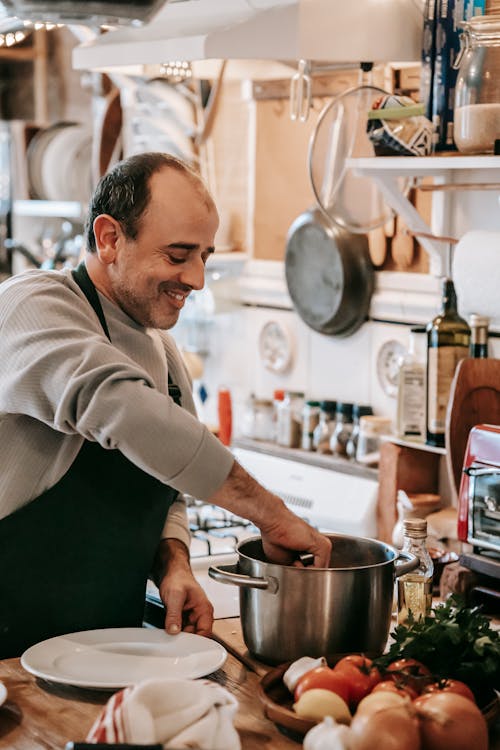 Happy middle aged ethnic male in casual clothes and apron smiling while putting dish from saucepan on plate in kitchen