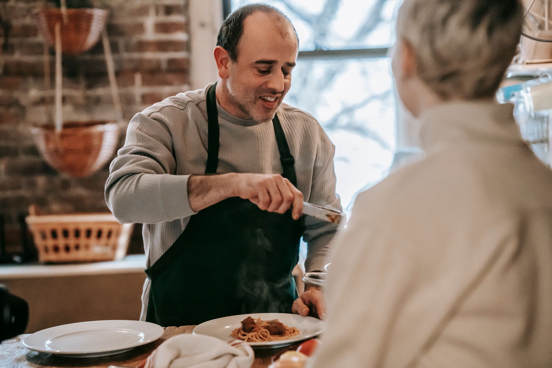 Happy middle aged ethnic mane in casual clothes and apron serving delicious homemade spaghetti with meatballs while standing at table in front of crop unrecognizable wife