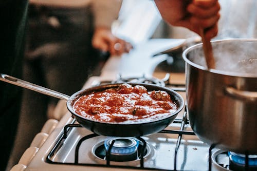 Free Crop unrecognizable person stirring boiling water in saucepan placed on gas stove near frying pan with appetizing meatballs in tomato sauce Stock Photo