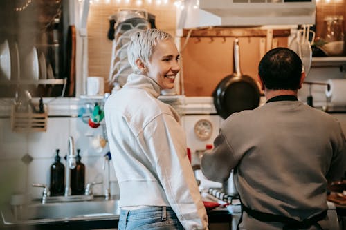 Side view of cheerful adult lady in casual clothes smiling while standing in kitchen near unrecognizable husband cooking food