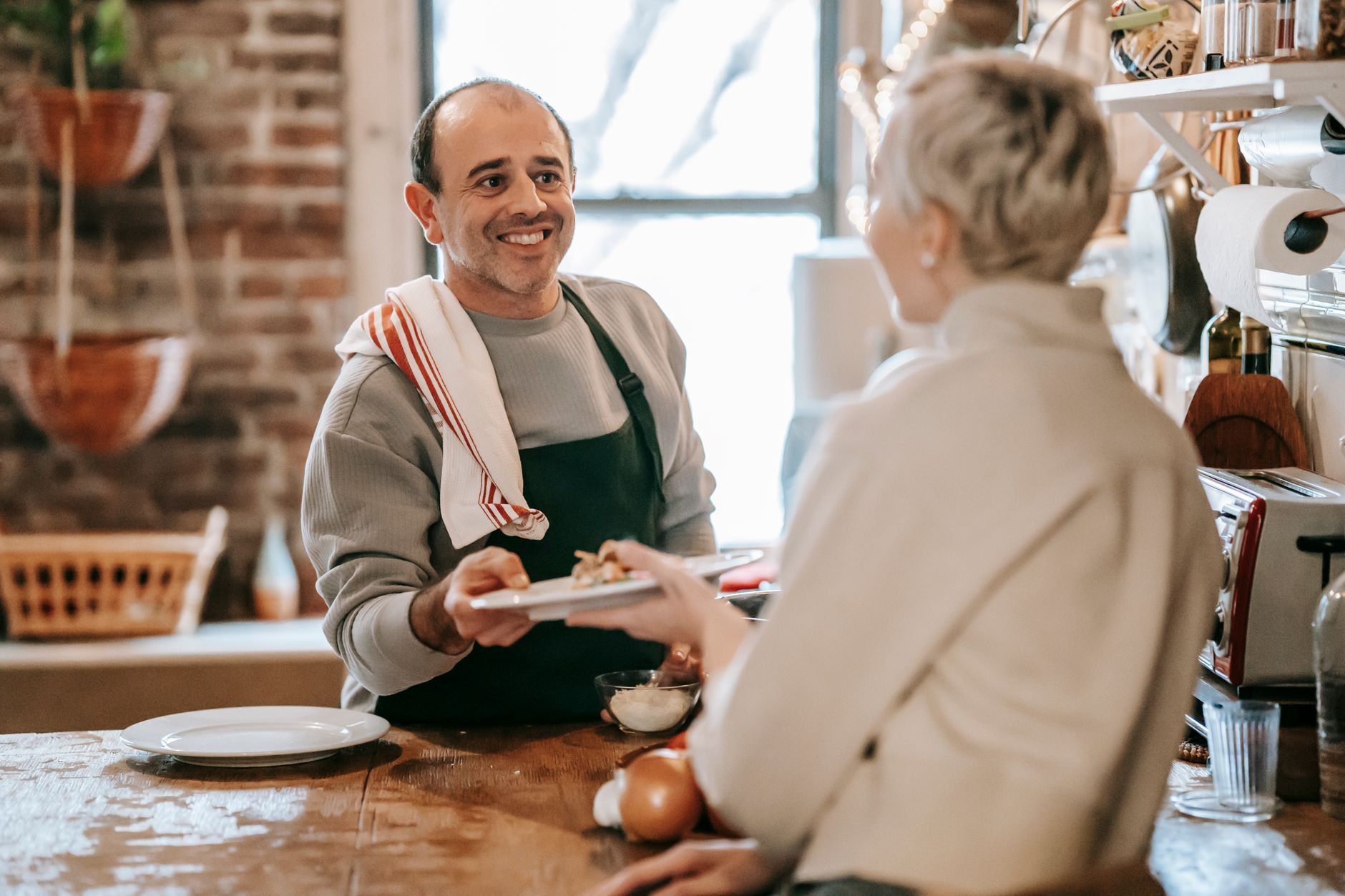 Happy adult ethnic man in casual clothes and apron giving plate with delicious homemade food to smiling wife sitting at table in kitchen