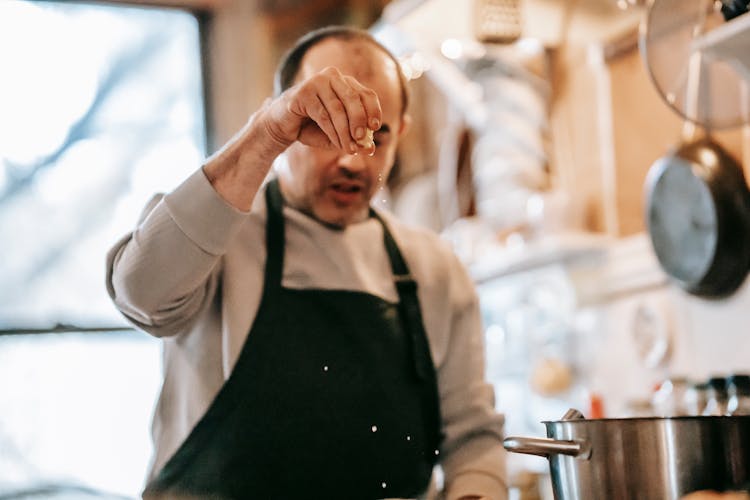 Serious Ethnic Man Adding Seasonings During Dinner Preparation In Kitchen