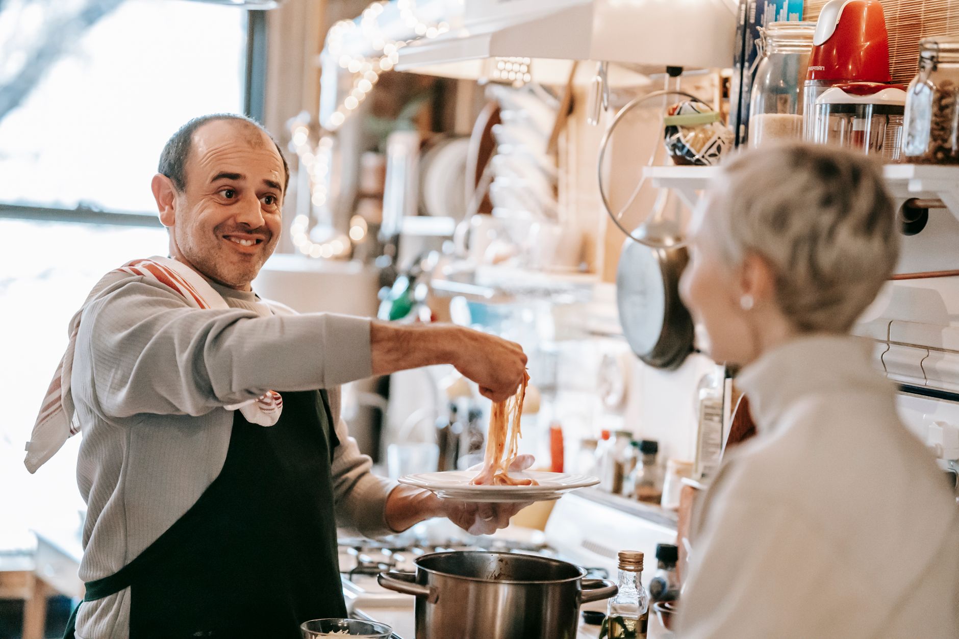Excited middle aged ethnic man in casual clothes and apron putting appetizing spaghetti on plate while chatting with wife during dinner in kitchen