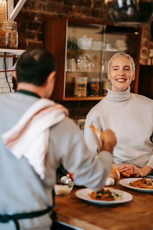 Free Happy adult female in casual clothes smiling while eating delicious spaghetti with tomato sauce with anonymous husband at home Stock Photo