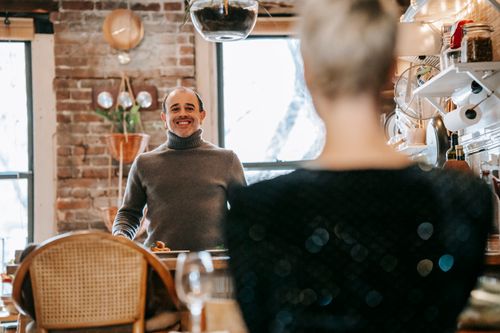 Free Cheerful middle aged man in stylish turtleneck smiling and looking at crop anonymous wife while standing at table in modern apartment Stock Photo