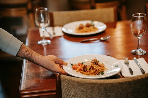 Unrecognizable waiter serving pasta on table with glasses in restaurant