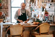 Positive middle aged male cooking food in bright room near counter with plates with pasta near onion and herbs near kitchenware and wine bottle near glasses and chairs near window